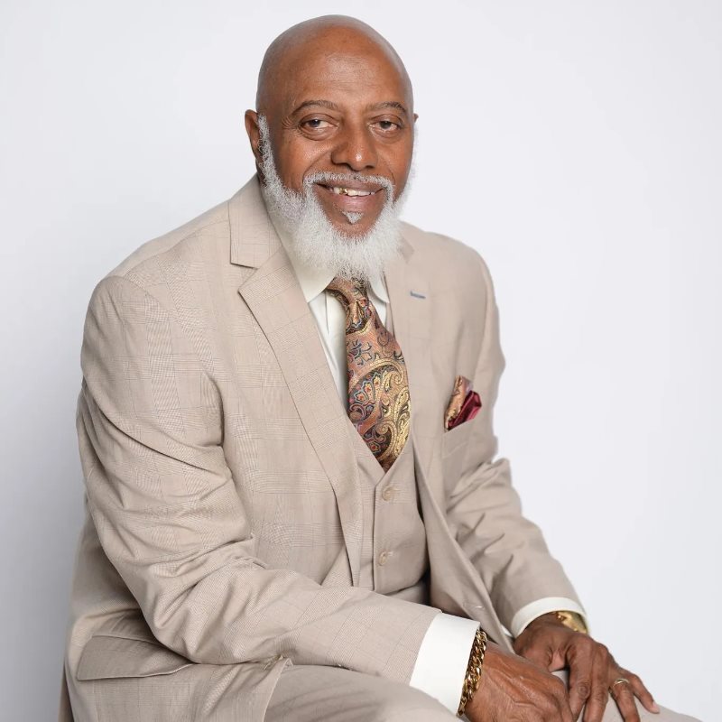 Bishop Rodney Gamble Sr., smiling, seated in a tan three-piece suit